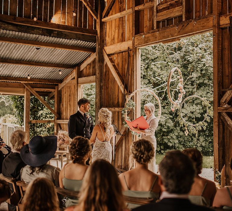 Hula hoop decor hangs at the altar behind the wedding celebrant with the bride in a boho lace wedding dress with the groom in traditional military outfit