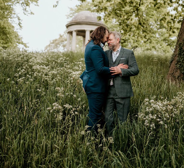 Groom in blue suit and groom in tartan suit embrace outdoors surrounded by lush greenery for botanical wedding 