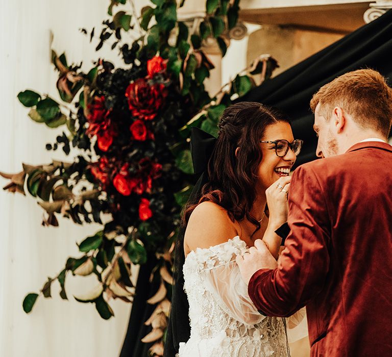 The bride and groom laugh together at the altar for their modern gothic wedding with black accents and burgundy details 