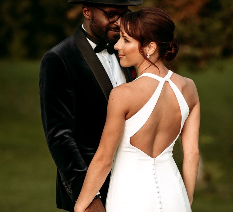 Black groom in a black velvet tuxedo jacket and black fedora hat accessory posing with the bride in an open back wedding dress 