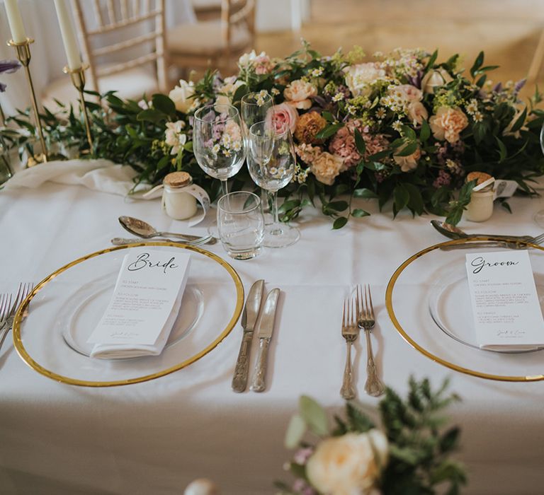 Gold rim clear charger plates with black and white wedding menu stationery on a white tablecloth with pastel wedding flower centrepiece 