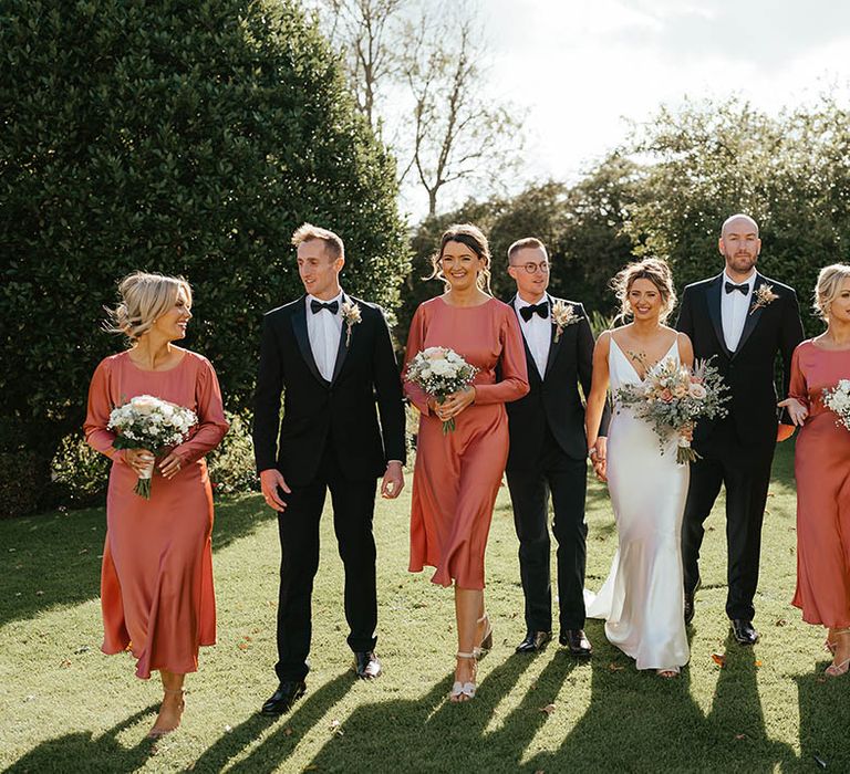 Bridesmaids in long sleeve satin midi bridesmaid dresses with the groom and groomsmen in black tie and the bride in a white satin bridal gown