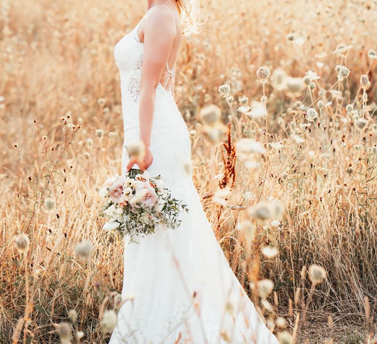 Smiling bride with her blonde hair in a half up half down hairstyle in a fitted lace wedding dress with low back holding a white bouquet 