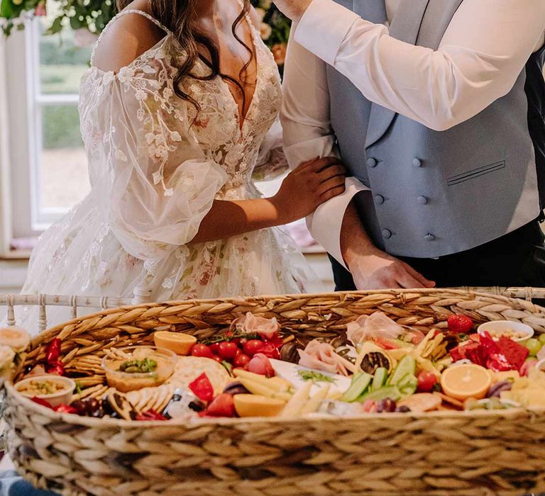 Groom in cornflower blue waistcoat and black suit trousers giving bride in v-neck long sleeve shoulder cut out 3d appliqué wedding dress with flecks of gold some wedding grazing platter 