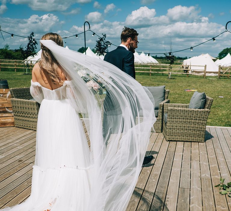 Bride in off the shoulder wedding dress with cathedral length veil walking with the groom in navy suit 