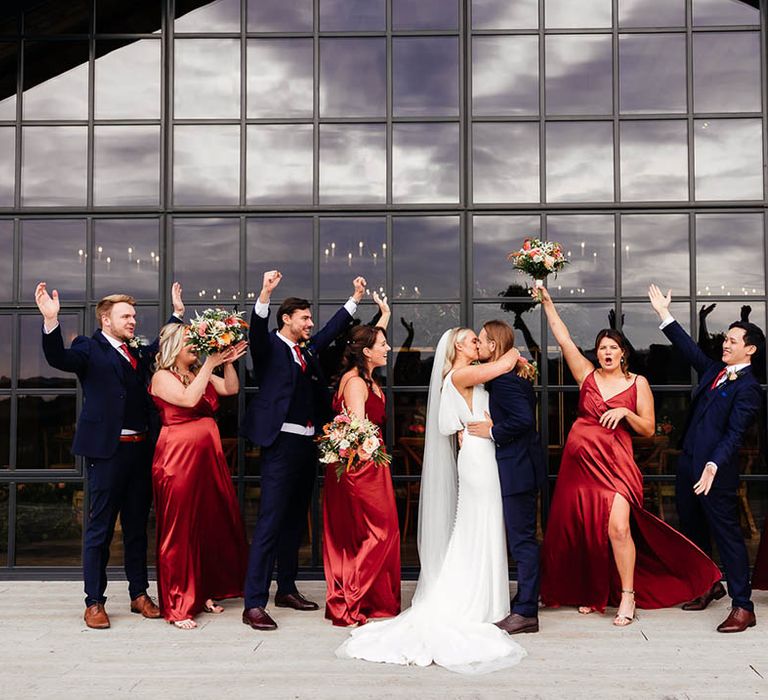 The bridesmaids in red satin bridesmaid dresses and the groomsmen in blue suits celebrate together as the bride and groom kiss