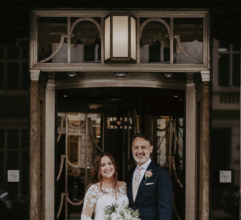 Groom in dark navy suit with floral tie and pink rose buttonhole posing with the pregnant bride at the entrance to the city wedding venue 