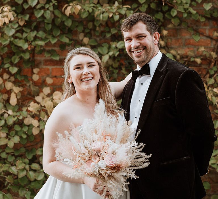 Bride holding dried faux wedding flowers in her bouquet with lace wedding dress with the groom in black velvet tux