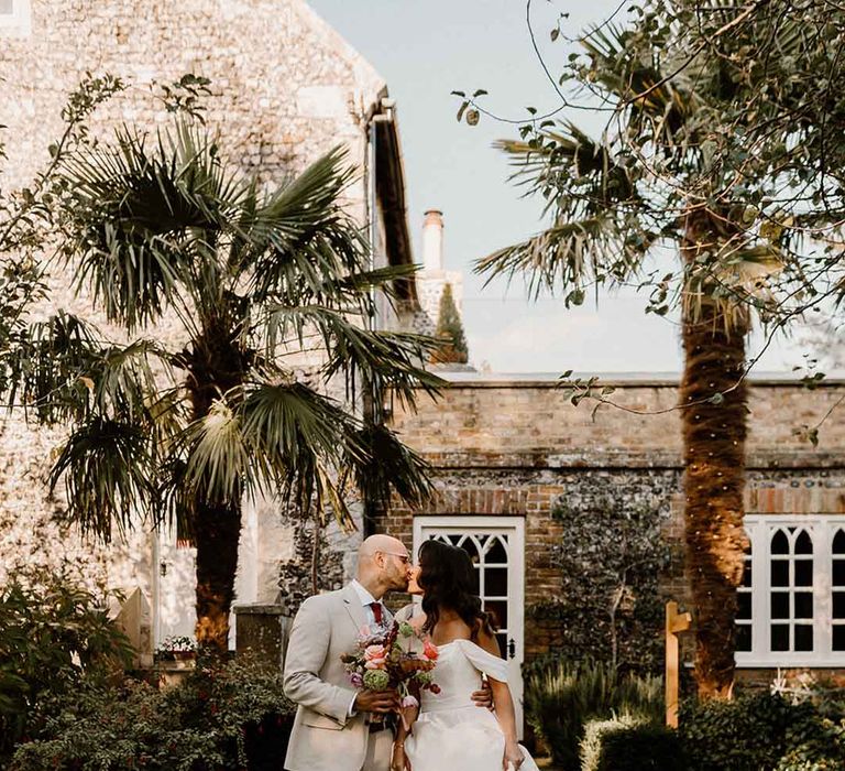 Groom in a stone suit kissing his bride in an off the shoulder wedding dress at Chapel House Estate wedding venue in Kent