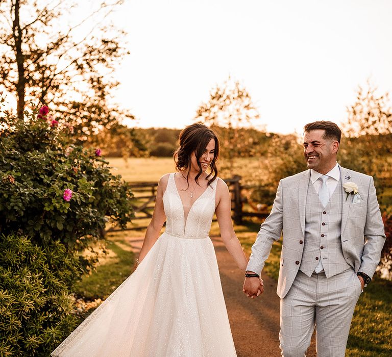 groom in a three-piece grey check suit holding hands with his bride in a sparkly wedding dress during golden hour 