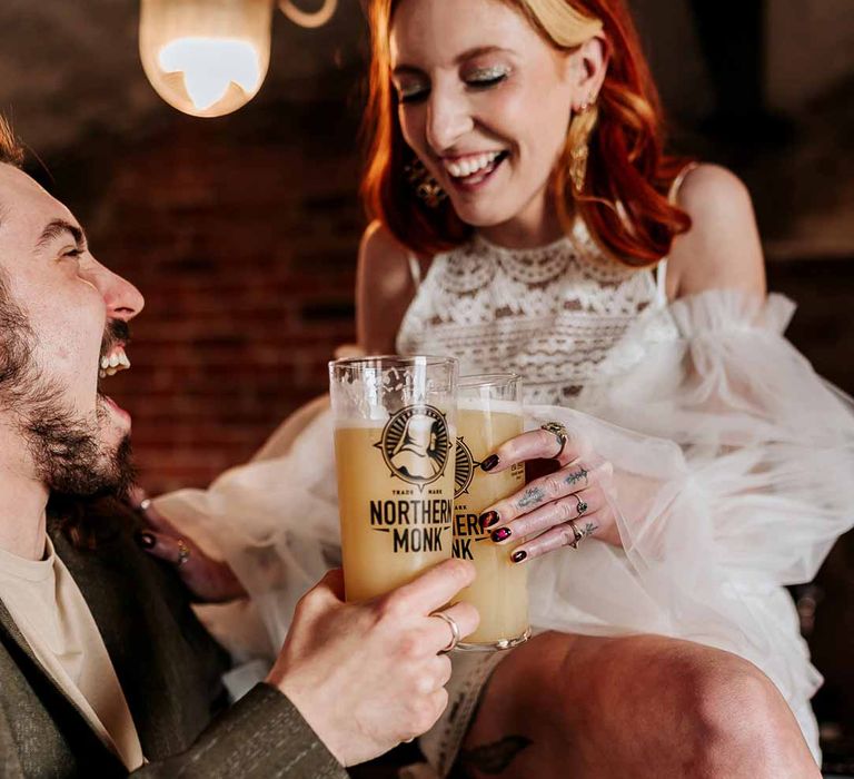 Bride in 70s-inspired boatneck short lace wedding dress with detachable tulle puff sleeves cheersing beer with groom in deep brown wedding suit and off-white t-shirt 