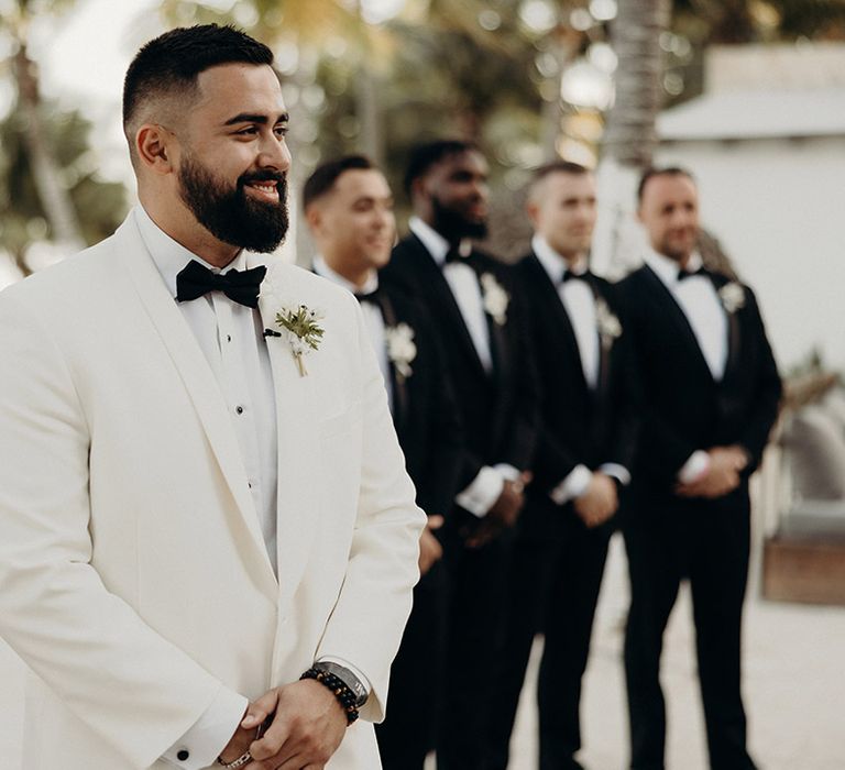 bearded groom in a white tuxedo jacket and black bow tie standing waiting for his bride