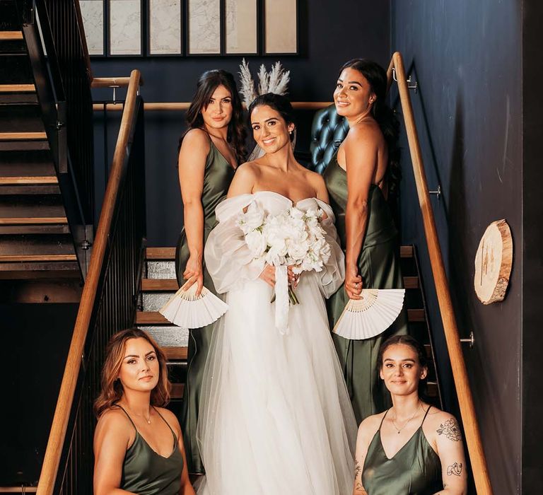 Bride in an off-the-shoulder wedding dress holds white rose wedding bouquet with bridesmaids in green stain bridesmaid dresses holding fan wedding favours at summer wedding