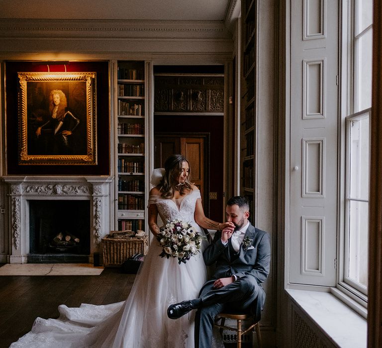 Groom sits on a chair with the bride standing next to him for couple portrait 