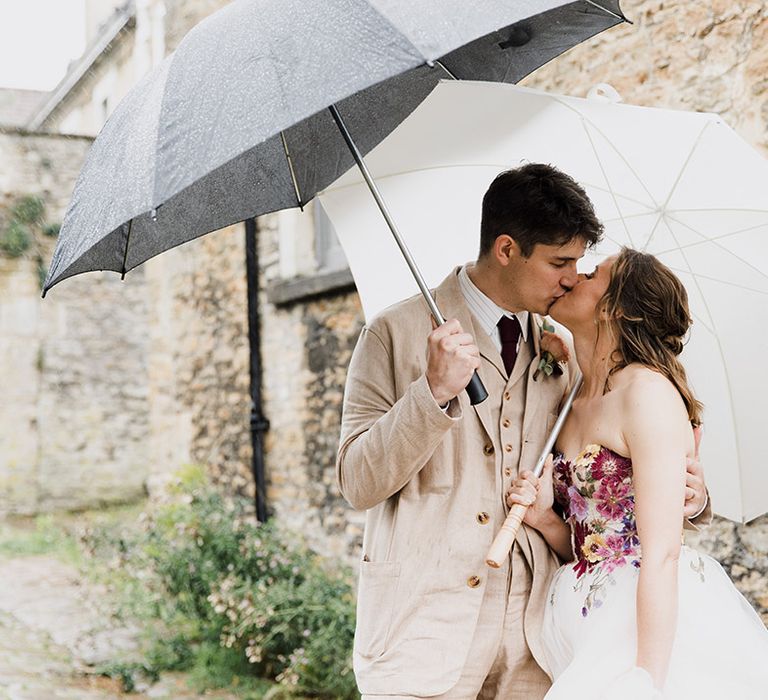 Bride in flower bodice wedding dress kissing groom in light beige suit under umbrellas on rainy wedding day 