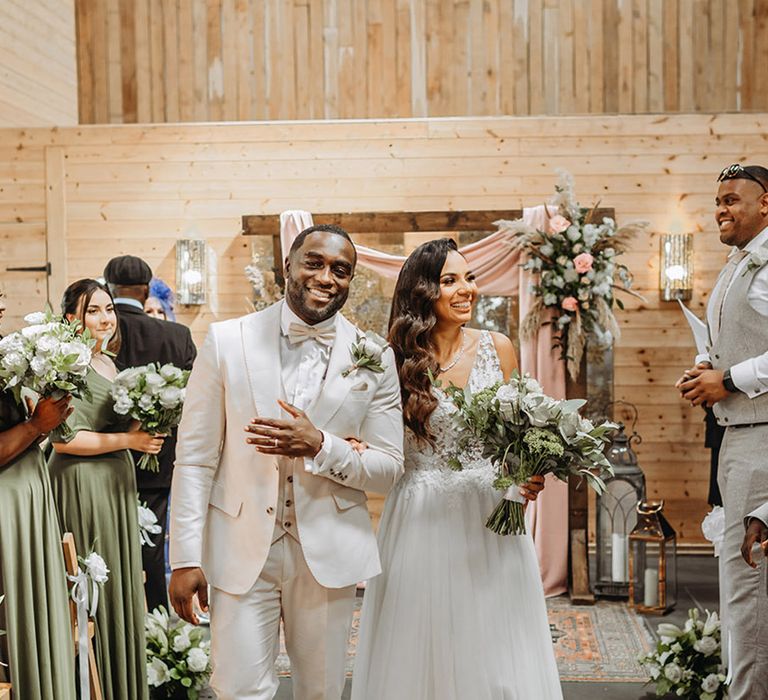 Bride in floral lace wedding dress walking with groom in three piece cream wedding suit back down the aisle as a married couple 