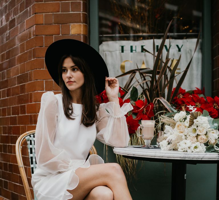 Bride sitting at a cafe in knee length wedding dress and fedora hat 