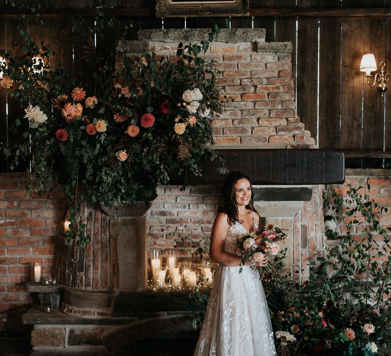 Bride in lace wedding dress standing in front of the fireplace decorated with flowers 