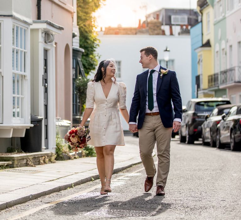 Bride in a Ba&sh registry wedding dress, birdcage veil and strappy shoes holding hands with her groom as they walk through London