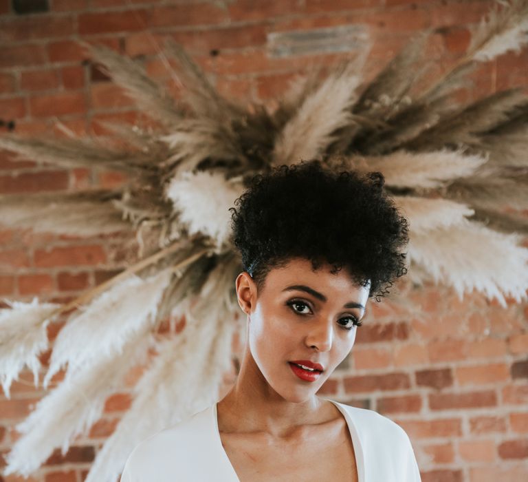 Wedding hairstyles for Black brides. A bride with a low cut dress has her hair cropped tightly round the sides and a mass of curls on top. She stands in front of a pampas grass installation.