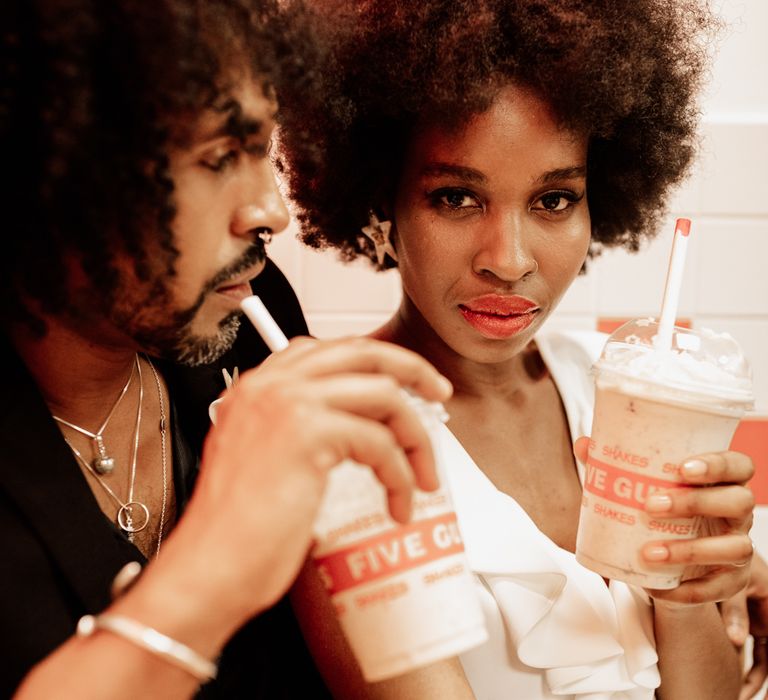 A bride and groon drink 5 guys shakes. The bride wears her hair in a natural afro with a central parting and looks straight to camera