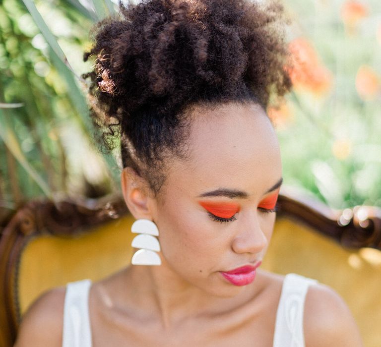 A bride with striking orange eyeshadow sits down. He curly hair is tied up high on her head.