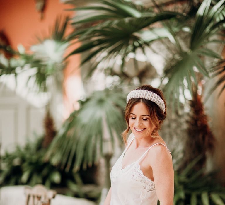 Smiling bride in white silk lace-trimmed playsuit and beaded bridal headband holding silver Jimmy Choo heeled sandals with white feather detail