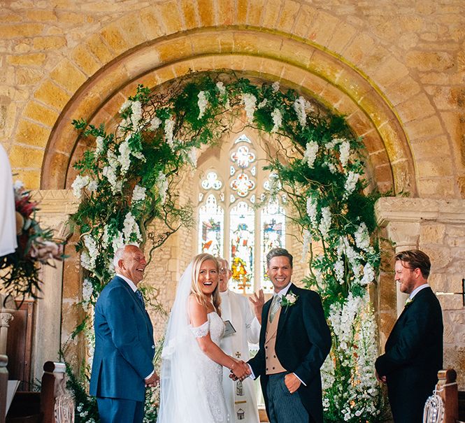 Bride and groom at Warwickshire church wedding with beautiful stained glass windows framed by a flower arch 