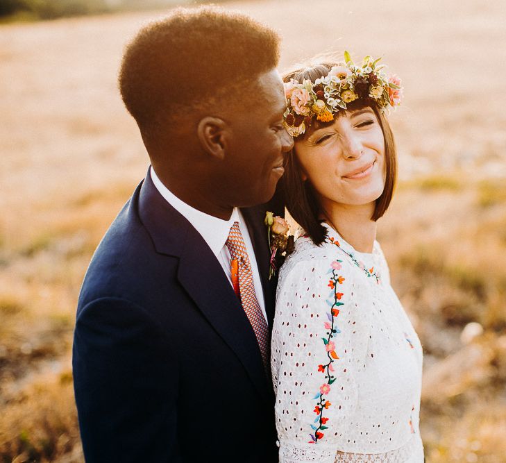 Golden hour portraits with groom in a navy suit smiling at his bride in a colourful flower crown 