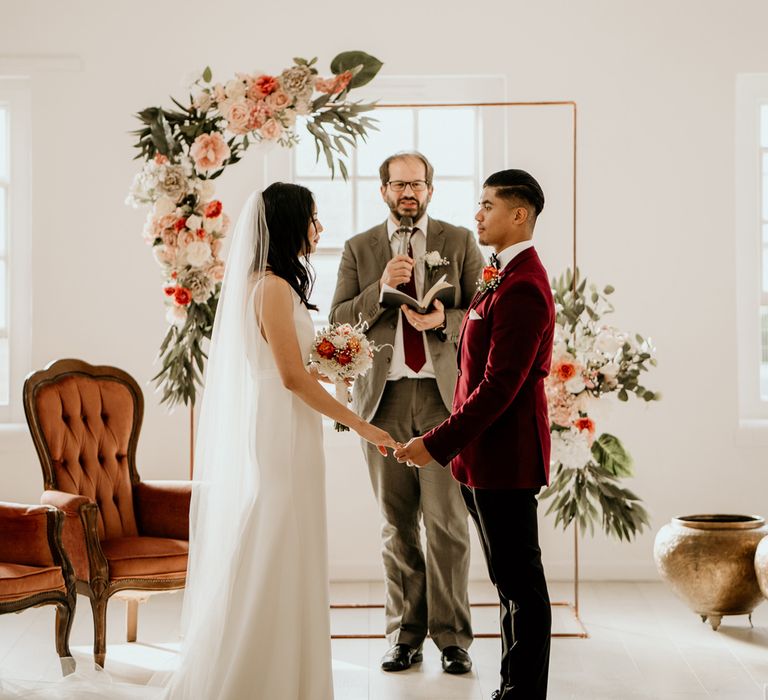 Bride in a minimalist Sarah Seven wedding dress exchanging vows with her groom in a burgundy jacket in front of a copper frame decorated with orange flowers