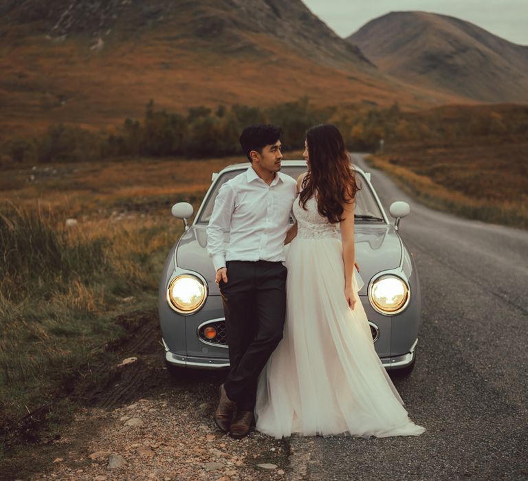 Couple stand in front of vintage car with headlights on in Scotland