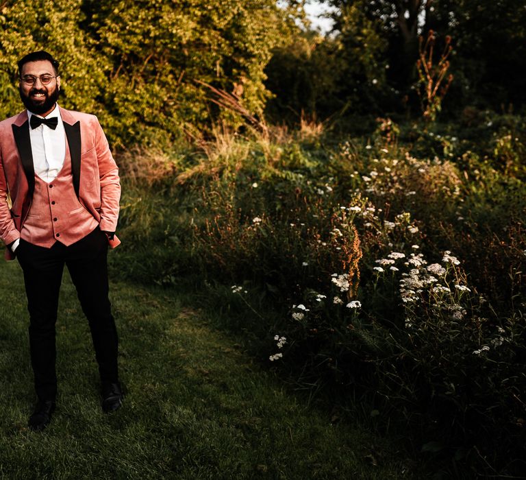 Groom stands in the garden and wears pink Topman suit 