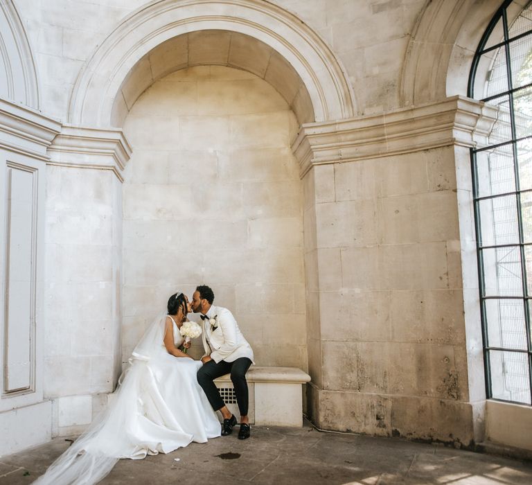 Ethiopian bride in a princess wedding dress, veil and tiara kissing her groom on a bench in a white tuxedo jacket 