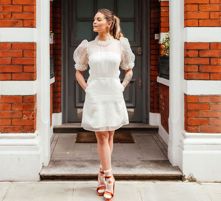 Stylish bride standing in a door wear in a knee length polka dot wedding dress with puff sleeves wearing platform sandals and floral socks 