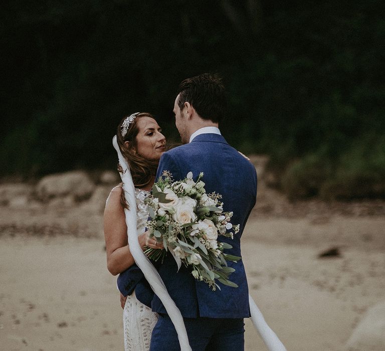 Bride & groom stand together on the beach as brides veil blows around them