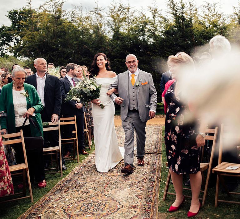 Bride in white strapless Vagabond wedding dress holding green and white botanical wedding bouquet walks down carpeted aisle arm-in-arm with father in grey suit and waistcoat with yellow tie and orange pocket square