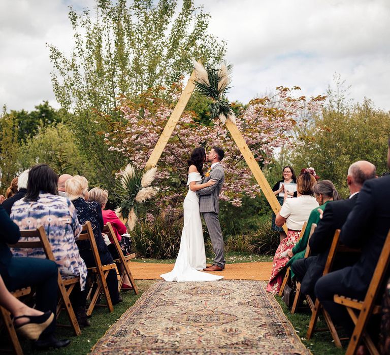 Bride in strapless Vagabond wedding dress and groom in grey Hugo Boss suit kiss under a wooden triangular wedding arch with pampas grass and palm leaf decor