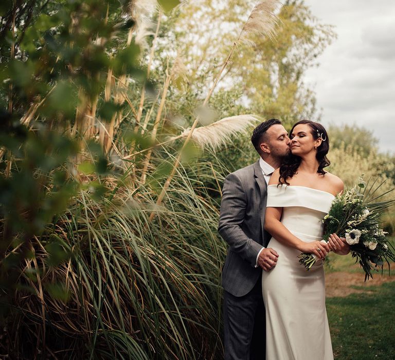 Bride in strapless Vagabond wedding dress holding white and green botanical bouquet stands outside by pampas grass being kissed by groom in grey suit at lakeside wedding