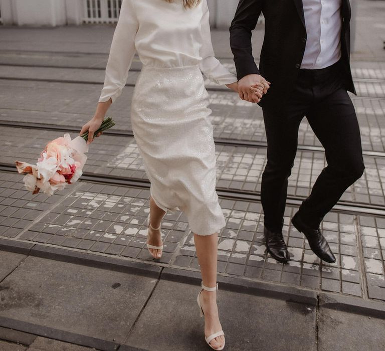 Bride and groom walking through the city, bride is wearing short mesh veil over her face with white, red and pink flowers on her head, and a long sleeve mid length wedding dress with sequin pencil skirt