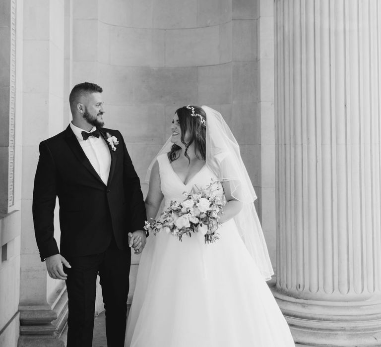 Bride & groom stand beneath white pillars on their wedding day for post-wedding photoshoot