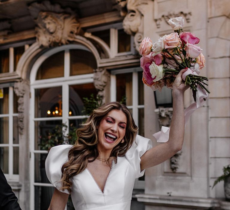 happy bride with long wavy hair, and pink eye shadow waving her blush pink, peeled back rose wedding bouquet in the air 