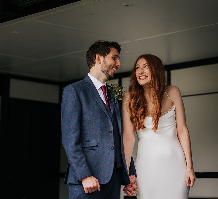 Smiley bride with red hair in a satin slip wedding dress holding hands with her husband in a navy check suit during their outdoor civil ceremony 