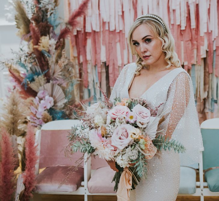 Bride in a sparkly dress wearing a pearl double headband holding a pink rose bouquet standing in front of a pastel tissue paper backdrop