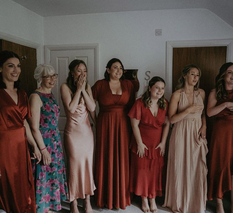 Bridesmaids in mismatched red and peach wedding dresses smile as they have first look at bride in her wedding dress before garden wedding with burnt orange wedding theme