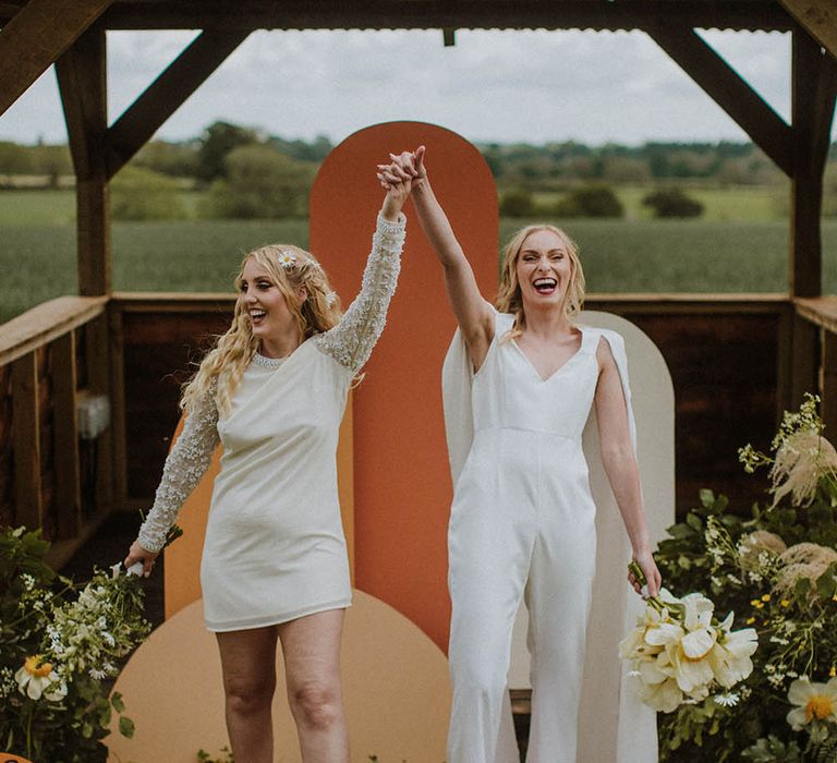 Sixties wedding with two brides in a mini wedding dress and bridal jumpsuit holding hands descending up the aisle 