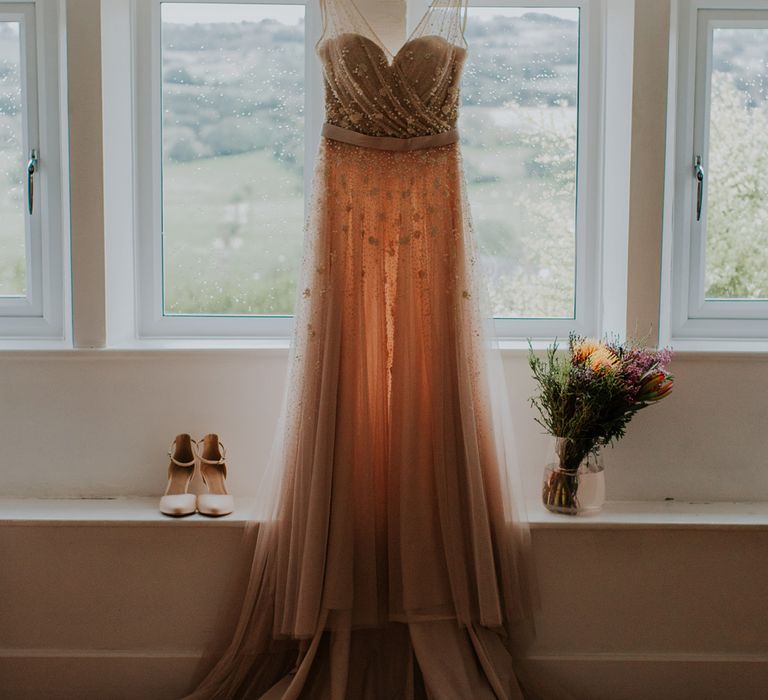 Pearl wedding dress with tulle and sequins hangs in front of window before barn wedding ceremony 