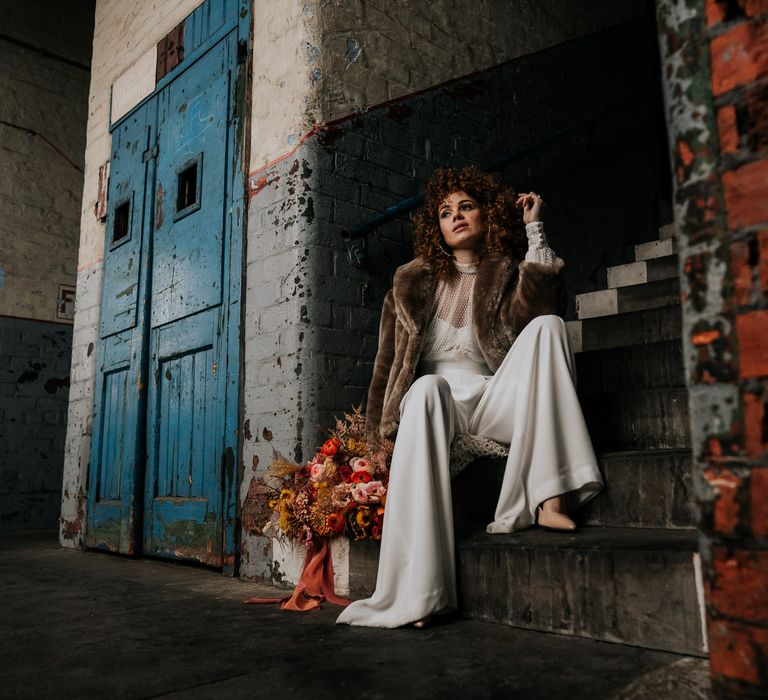 Bride in a wedding jumpsuit and brown faux fur coat sitting on a step in an industrial wedding venue with an autumn wedding bouquet 