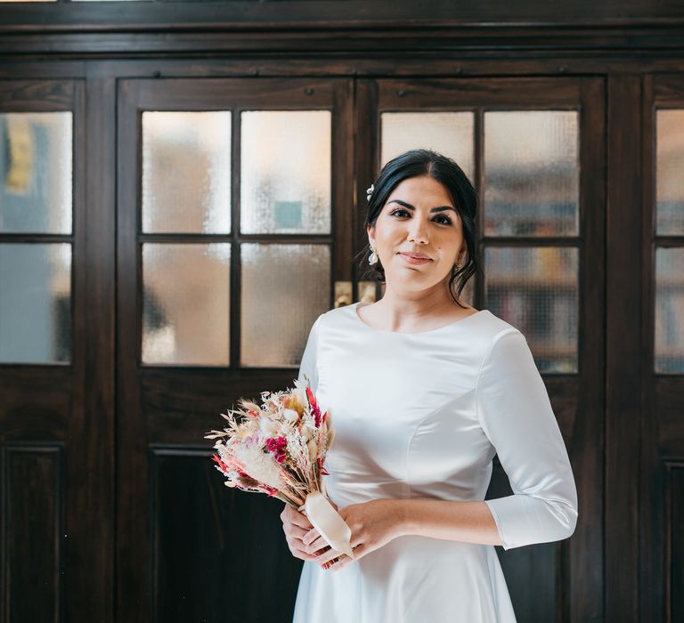 Bride smiles as she holds dried flower bouquet and wears elegant wedding gown with her dark hair pulled back