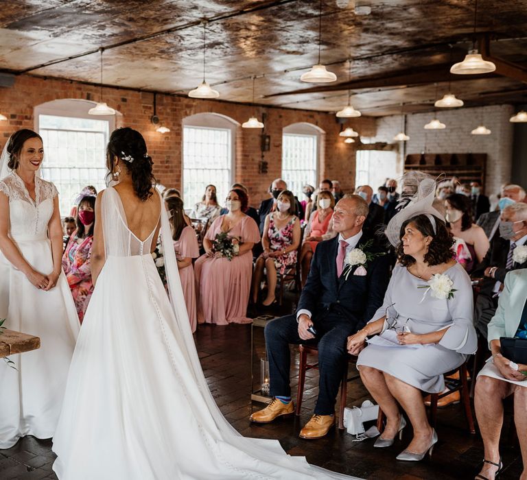 Bride in low mesh back wedding dress and shoulder veil details stands with bride in lace capped sleeved wedding dress and veil in front of guests during wedding ceremony at The West Mill Derby