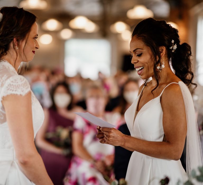 Bride with curled ponytail and white cami wedding dress smiles whilst reading wedding vows to bride in lace capped sleeved wedding dress and veil in front of guests at The West Mill Derby during wedding ceremony